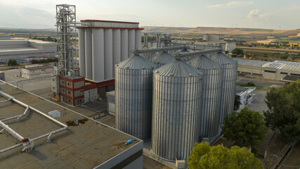 Aerial view of a massive industrial grain facility featuring towering silver metal silos and white storage bins. The complex includes elevator towers and conveyors set against a rural landscape. © Stefano Tammaro