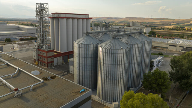 Aerial view of a massive industrial grain facility featuring towering silver metal silos and white storage bins. The complex includes elevator towers and conveyors set against a rural landscape.
