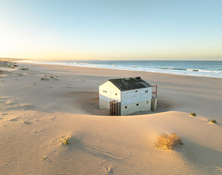 Costa da Caparica, Portugal - 29 January 2025: Aerial view of a weathered building, embraced by golden sands, whispers tales of the ocean's relentless caress.