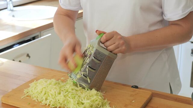 Woman is cutting up a head of cabbage on a cutting board. She is using a grater to shred the cabbage