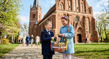 Dwoje dzieci w odświętnych płaszczach trzymają udekorowany wiklinowy koszyk ze święconką przed kościołem