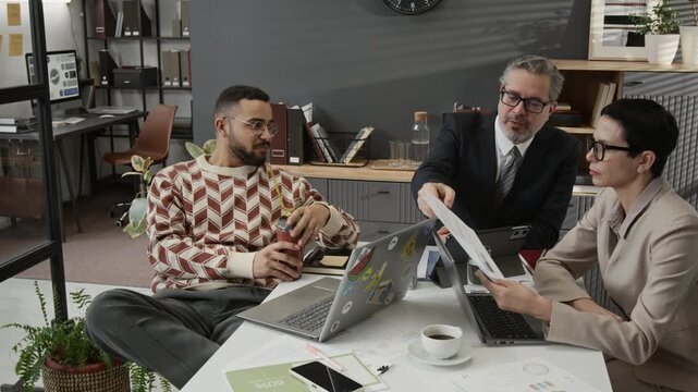 Positive zoomer drinking from travel mug and discussing document given to him by smiling businessman while having office meeting with middle-aged colleagues