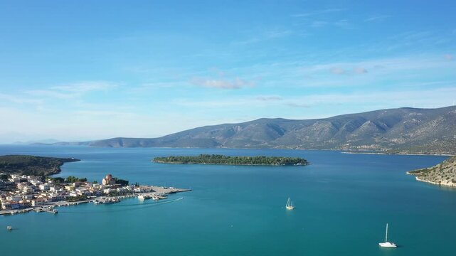 Aerial view of the tranquil bay and coastal village of Kilada, Greece, with clear blue water, a small island, and distant mountains under a bright sky.