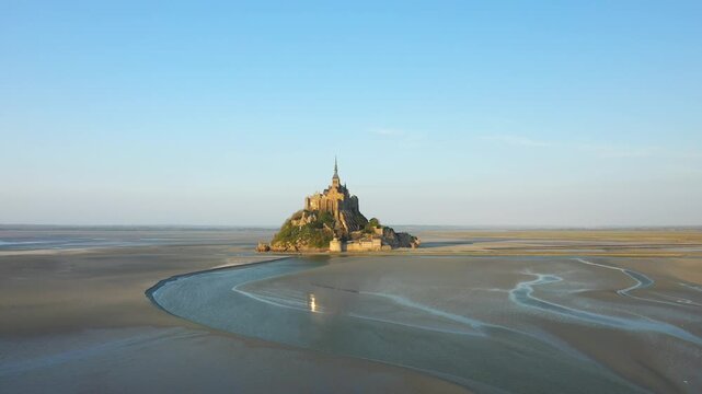 Aerial view of Mont Saint Michel surrounded by tidal flats under clear blue sky in Normandy, France. The iconic island abbey stands isolated amidst expansive sand and water patterns.