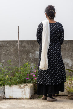 Back view of a woman in traditional black patterned attire and white dupatta, standing amidst plants in a rooftop garden in Bangladesh. Authentic South Asian urban gardening and lifestyle