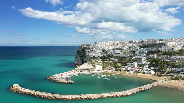 Scenic aerial view of the whitewashed buildings of Peschici perched on a cliff above the turquoise Adriatic Sea, with a marina and boats in the harbor below. Picturesque coastal landscape under a