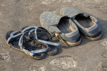 A pair of worn-out blue female strappy sandals and men's canvas slip-on shoes on dusty concrete ground. A poignant concept of hardship, poverty, and used footwear in a rugged outdoor setting