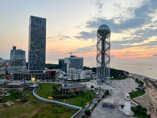 Obraz premium Aerial view of Batumi, Georgia showcasing modern skyscrapers, the Alphabet Tower, and the Black Sea coastline during sunset with vibrant clouds in the sky