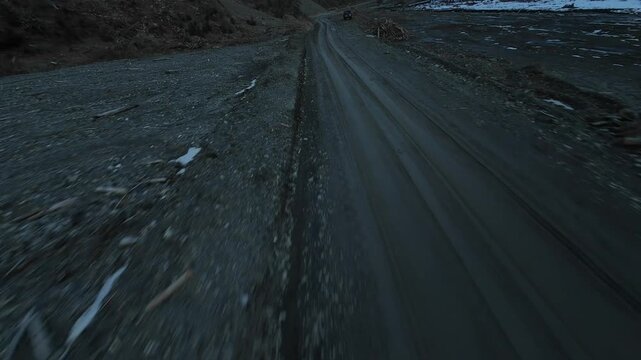 Fast low-altitude FPV drone flight skimming over a dark gravel mountain road with patches of snow, dead wood debris, and a moody, desolate winter landscape.