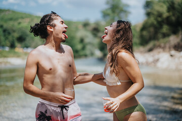 Two friends in swimwear laugh and stick out their tongues by a river. A sunny day outdoors captures...