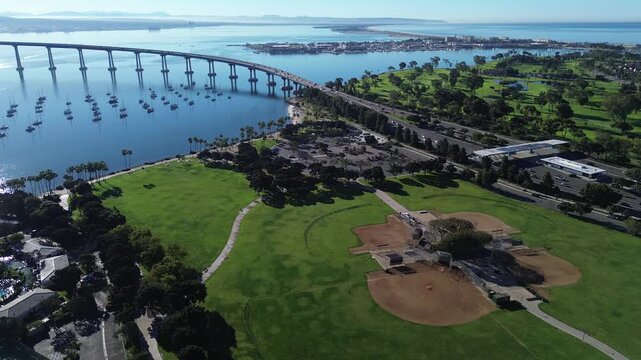Park fields and bridge shows Tidelands Park ballfields near Mullinex Dr with Coronado Bridge rising over anchored boats. Sunlit grass, clean rooflines, shimmering bay textures define Coronado, CA