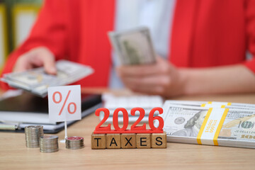 On a wooden desk, a person sorts cash while looking at a calculator, preparing for taxes due in...