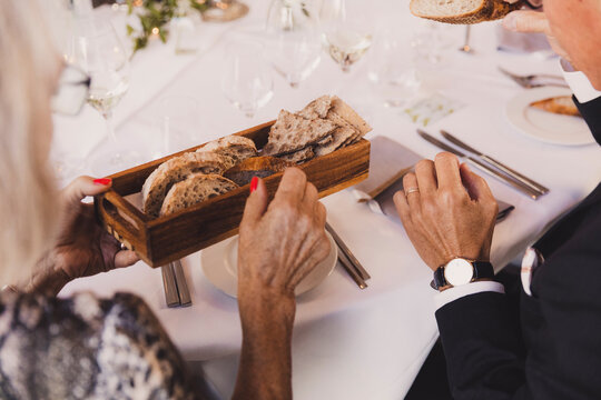 Elegant senior couple sharing a wooden bread basket during a formal dinner party or wedding reception. Close-up of hands with red nails and a luxury watch at a white tablecloth restaurant setting.