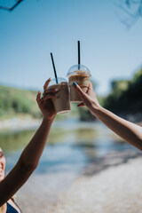 Two friends raise iced coffee cups in a bright lakeside scene. Outdoor summer moment of friendship...