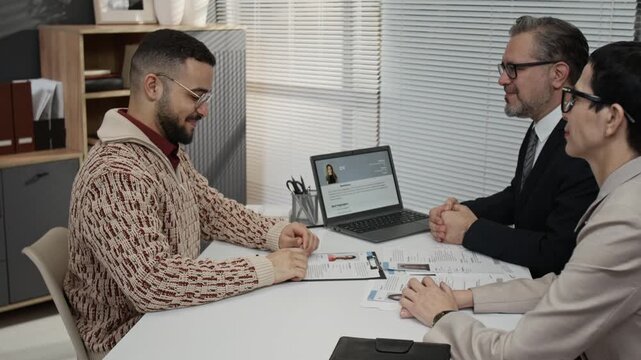 Zoom out side view of smiling gen Z man sealing deal with handshakes after job recruitment meeting with two mature HR specialists sitting at office table in front of him