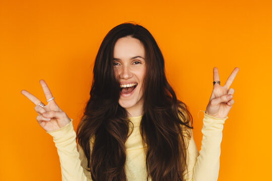 Portrait of playful young woman showing peace sign gesture on orange background, happy girl with long hair making V-sign and smiling at camera, positive vibes and youthful lifestyle concept, wow