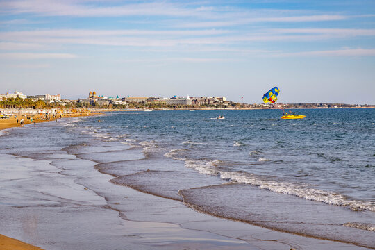 Wide view of a quiet sandy coastline with soft waves. Bright yellow boat with a parasailer in the blue sky above the sea during a sunny spring day in Evrenseki, Side, Turkey.