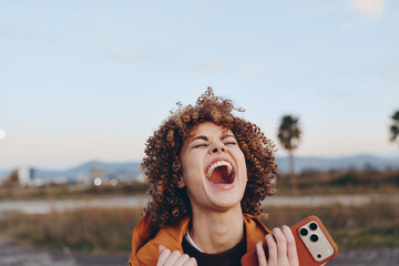 A happy woman with curly hair wearing a rainbow sweater smiles widely while holding a smartphone...