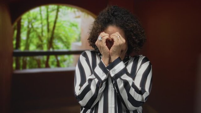 Woman with curly hair in striped blouse forming heart shape with her hands in a building corridor with arch window; love.