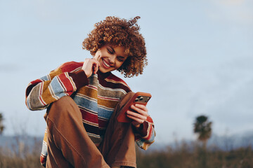 Smiling woman with curly hair wearing a rainbow sweater sits outdoors using a smartphone, enjoying...