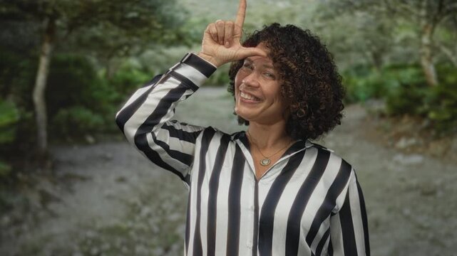 Woman makes l sign with her hand to forehead in forest, smiling in black and white striped shirt with pendant necklace; playful teasing.