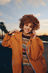 Smiling woman with curly hair wearing rainbow sweater and orange jacket talks on smartphone...