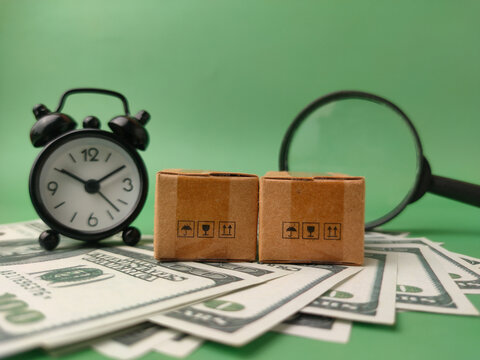 Two miniature cardboard shipping boxes placed on a fan of US dollar bills, accompanied by a small black alarm clock and a magnifying glass in the background
