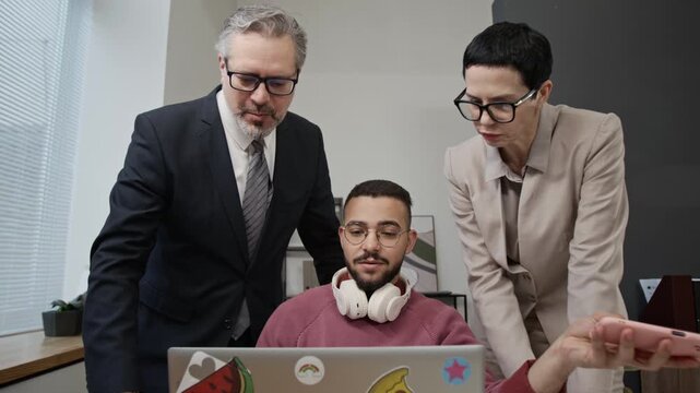 Two formally dressed middle-aged managers speaking with gen Z man and pointing at screen of his laptop while checking his work in office