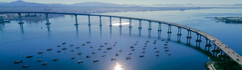 Panorama aerial curved bridge overlook frames busy Coronado Bridge above anchored boats near sandy edge of Tidelands Park and Glorietta Bay. Sunlit water patterns, palm silhouettes, shoreline, CA © trongnguyen