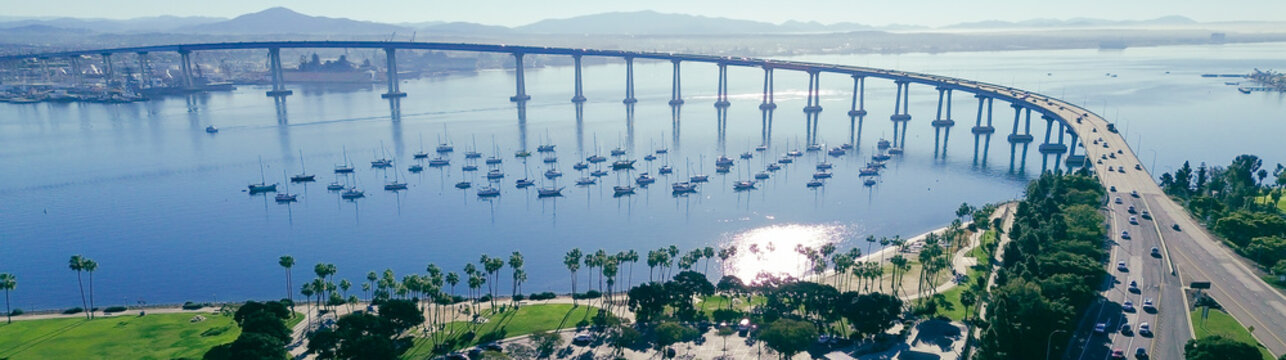 Panorama aerial park and roundabout layout with anchored bay corridor Coronado Bridge rising over San Diego Bay, moored sailboats and palm lined shoreline near Tidelands Park. Reflective water, CA