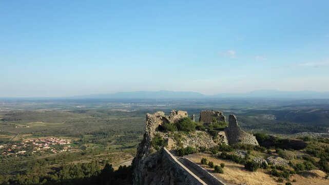 Wide view of the stone ruins of Chateau de Opoul Perillos overlooking the rural landscape and village below under a clear blue sky in southern France.