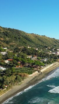Aerial view of the scenic coastline and hillside in Acciaroli, Italy, with turquoise waves and lush greenery under a clear blue sky.
