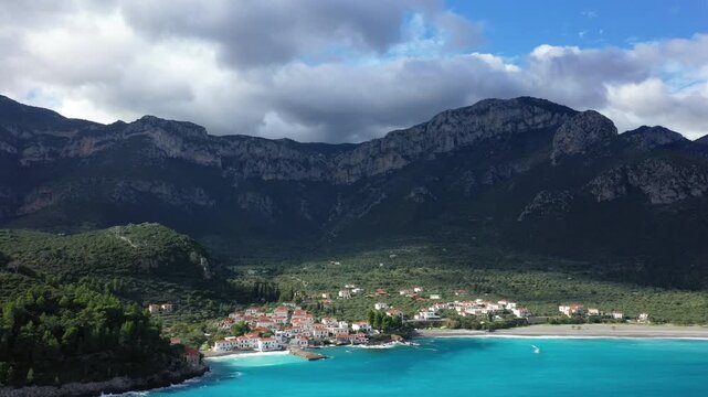 Aerial view of a picturesque seaside village with turquoise waters and dramatic mountains near Monemvasia, Greece. Lush greenery and traditional white houses line the coastline under a partly cloudy
