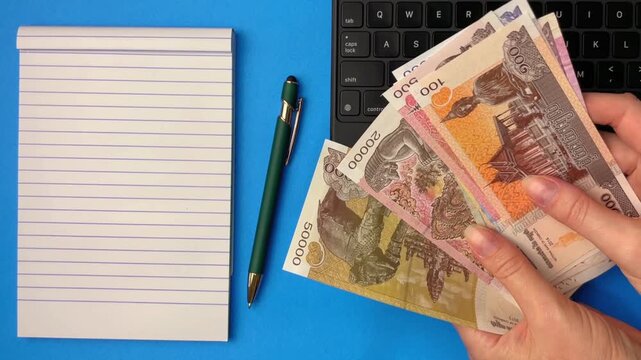 A woman's hands count Cambodian banknotes next to a notepad and pen. Concepts for tracking expenses, budgeting, financial control, and spending planning.