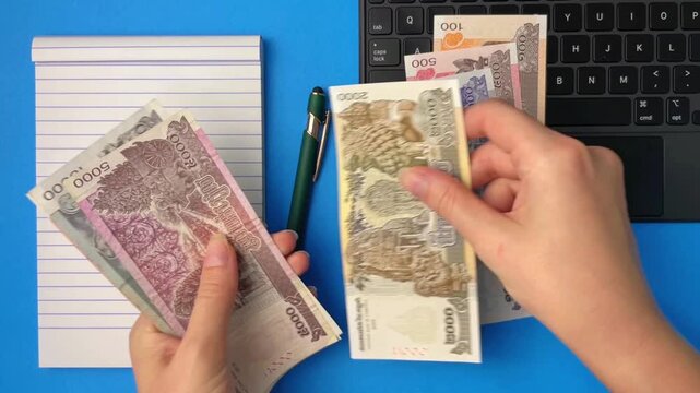 An overhead view of hands counting Cambodian currency next to a laptop and notepad. Conceptual concepts of remote work, online earnings, the digital economy, and income management.