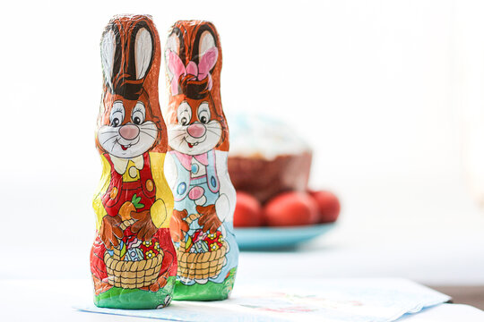 Two festive Easter chocolate bunnies in foil wrappers with blurred Easter cake and red eggs on background.
