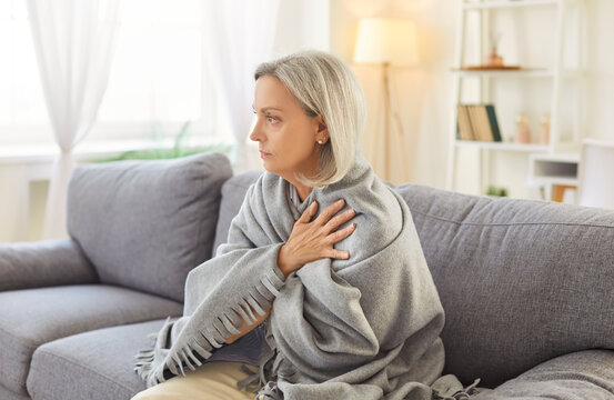 Portrait of senior gray-haired woman sitting on sofa in living room at home wrapped in cozy blanket trying to keep warm. Elderly retired person freezing in winter in chilly room with low temperature.