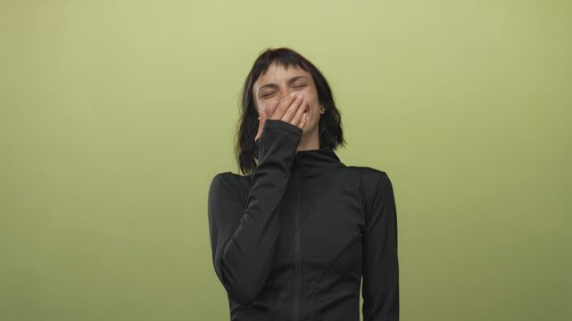 Young hispanic woman wearing black zip up jacket and active top with hand covering mouth laughing in studio; joy.
