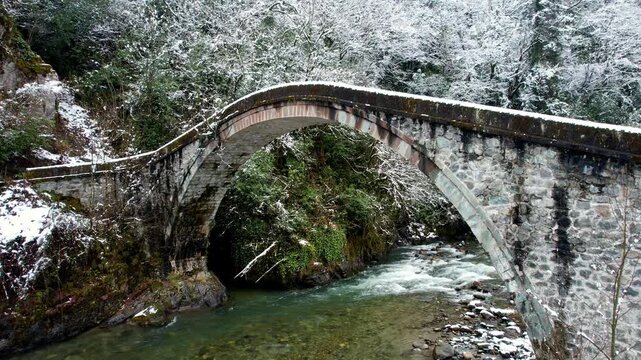 Gentle snowfall over a historic stone arch bridge crossing a clear mountain stream in Ayder Plateau, Rize, Turkey. Snow-covered forest and flowing water create a peaceful winter nature landscape.