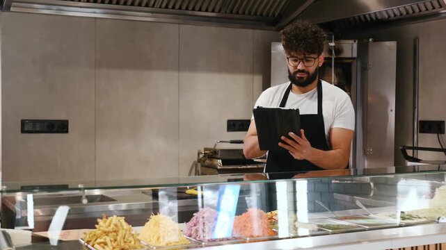 Male worker in a black apron checks a tablet in a modern kitchen, with food preparation items and equipment visible in the background