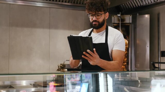 Male worker in a black apron checks a tablet in a modern kitchen, with food preparation items and equipment visible in the background