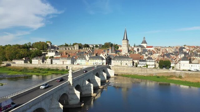 A scenic view of the historic stone bridge crossing the Loire River and the picturesque town of La Charite sur Loire under a clear blue sky. Traditional French architecture and church spires dominate