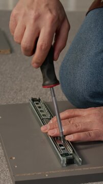 Vertical close-up of hands of unrecognizable man joining runner mechanism to drawer panel with bolts and screwdriver, assembling chipboard knockdown cabinet at home