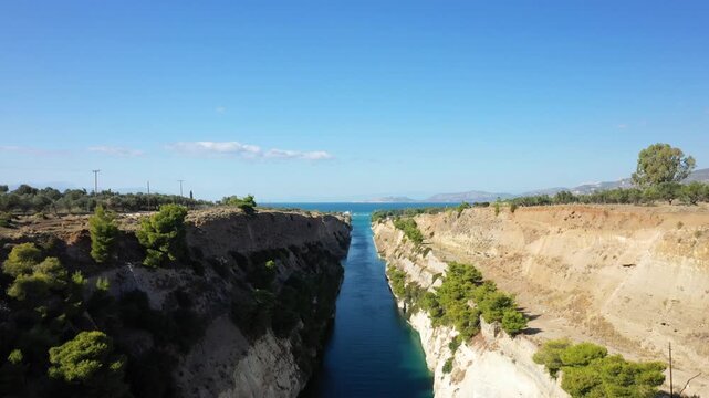 Aerial view of the Corinth Canal cutting through rocky terrain with blue sea and distant mountains under a clear sky in Greece.