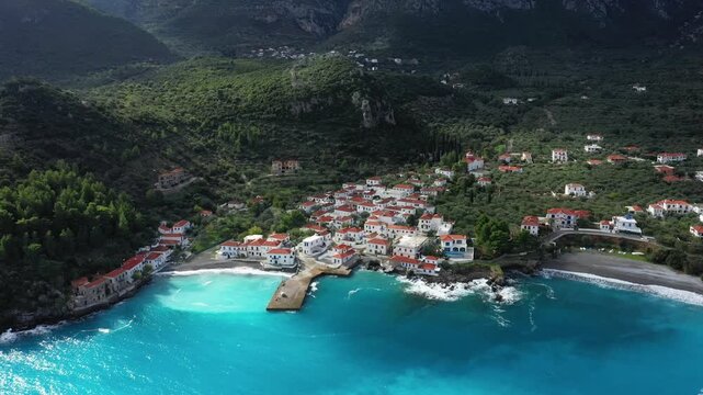 Drone shot of a picturesque coastal village with red roofed houses, turquoise sea, and lush green hills near Monemvasia, Greece.