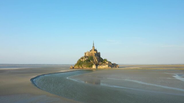 Aerial view of Mont Saint Michel surrounded by tidal flats under a clear blue sky in Normandy, France. The iconic abbey rises above the sandy landscape, showcasing its historic architecture.