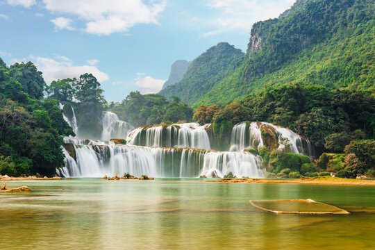 Ban Gioc waterfall fans across a wide rocky ledge into a golden-green river, jungle-covered karst cliffs rising on both sides under blue sky.