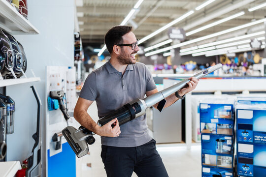 A cheerful male retail employee has fun while working by pretending a cordless vacuum cleaner is an electric guitar in a modern home appliance showroom.