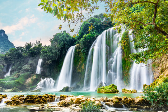 Ban Gioc waterfalls tumble down stepped rock faces draped in green moss, mist rising over a calm jade pool surrounded by dense rainforest.