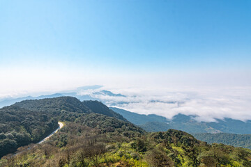 Fototapeta premium A winding mountain road curves through forested green hills blanketed in low cloud, blue sky and sunlight breaking above the misty ridgeline.